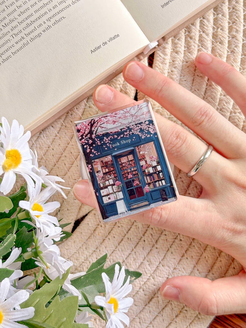 May include: A clear, rectangular phone grip featuring a miniature illustration of a blue bookstore with the text "Book Shop" and pink cherry blossoms. The grip is held against a neutral background.