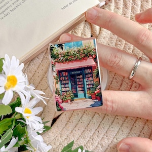 May include: A rectangular phone grip featuring a colorful illustration of a quaint bookstore with a pink awning and potted flowers. The bookstore has a blue door and is filled with bookshelves. The text "BOOKSHOP" is visible.
