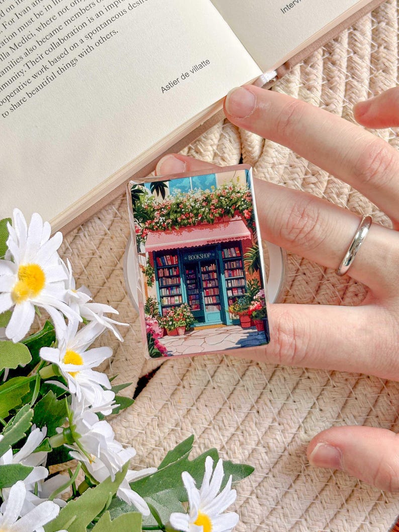 May include: A rectangular phone grip featuring a colorful illustration of a quaint bookstore with a pink awning and potted flowers. The bookstore has a blue door and is filled with bookshelves. The text "BOOKSHOP" is visible.