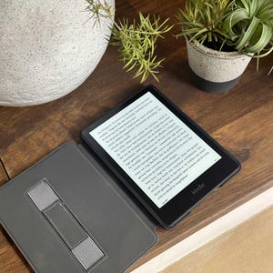 May include: A black Kindle e-reader with a gray protective cover featuring an elastic hand strap. The screen displays Dutch text, showing a paragraph from a book. The device has a black bezel with the Kindle logo printed at the bottom center.