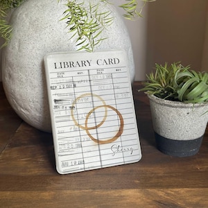 May include: A white library card-themed coaster with the text "LIBRARY CARD" and coffee ring stains. The coaster has a vintage design with dates and the name "Sherry" written on it. The coaster is displayed on a wooden surface.