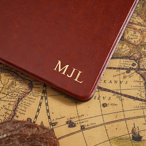 May include: A close-up of a brown leather notebook with the gold initials "MJL" embossed on the cover. The notebook is resting on a vintage map, with a dark brown rock in the foreground.