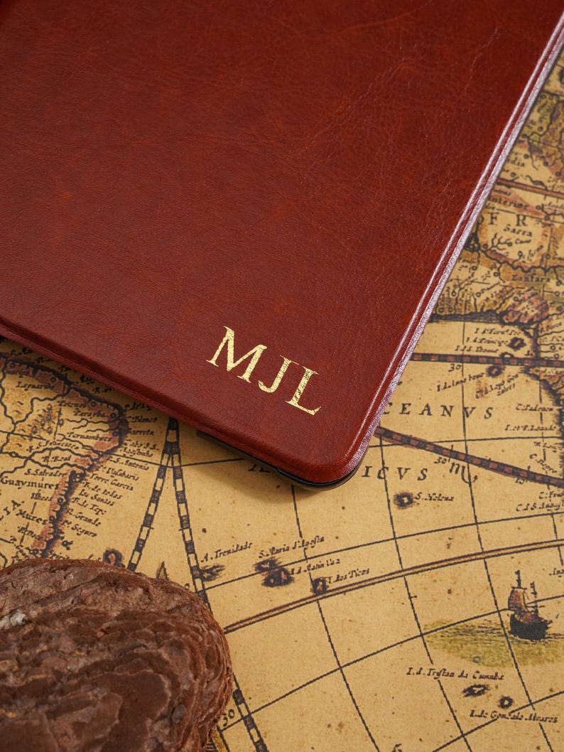 May include: A close-up of a brown leather notebook with the gold initials "MJL" embossed on the cover. The notebook is resting on a vintage map, with a dark brown rock in the foreground.