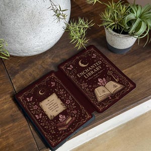May include: A dark red e-reader case featuring a gold-toned illustration of an open book, crystals, butterflies, and a crescent moon. The text reads "ENCHANTED LIBRARY" and "Some stories whisper. Some sparkle. Some change everything."
