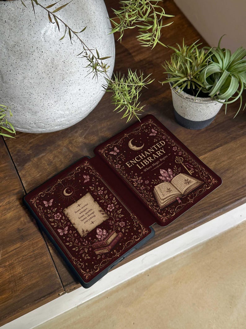 May include: A dark red e-reader case featuring a gold-toned illustration of an open book, crystals, butterflies, and a crescent moon. The text reads "ENCHANTED LIBRARY" and "Some stories whisper. Some sparkle. Some change everything."