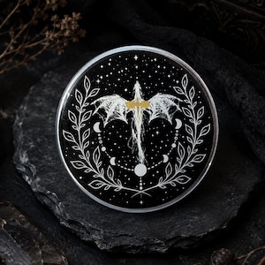 Puede incluir: Un posavasos circular negro con una ilustración de líneas blancas de un dragón con las alas extendidas, rodeado por un ciclo de fases lunares y dos ramas frondosas simétricas. El pecho del dragón tiene un pequeño detalle de color dorado.
