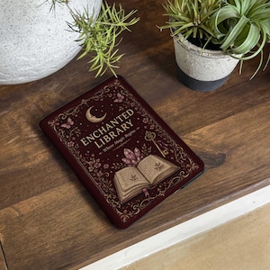 May include: A dark red e-reader cover featuring a gold-colored illustration of an open book, crystals, a key, butterflies, and a crescent moon. The text reads ENCHANTED LIBRARY Where Magic Begins in gold lettering against a decorative border.