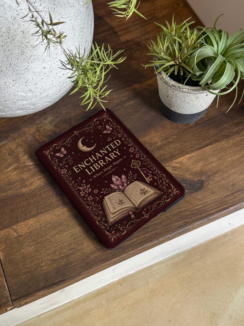 May include: A dark red e-reader cover featuring a gold-colored illustration of an open book, crystals, a key, butterflies, and a crescent moon. The text reads ENCHANTED LIBRARY Where Magic Begins in gold lettering against a decorative border.