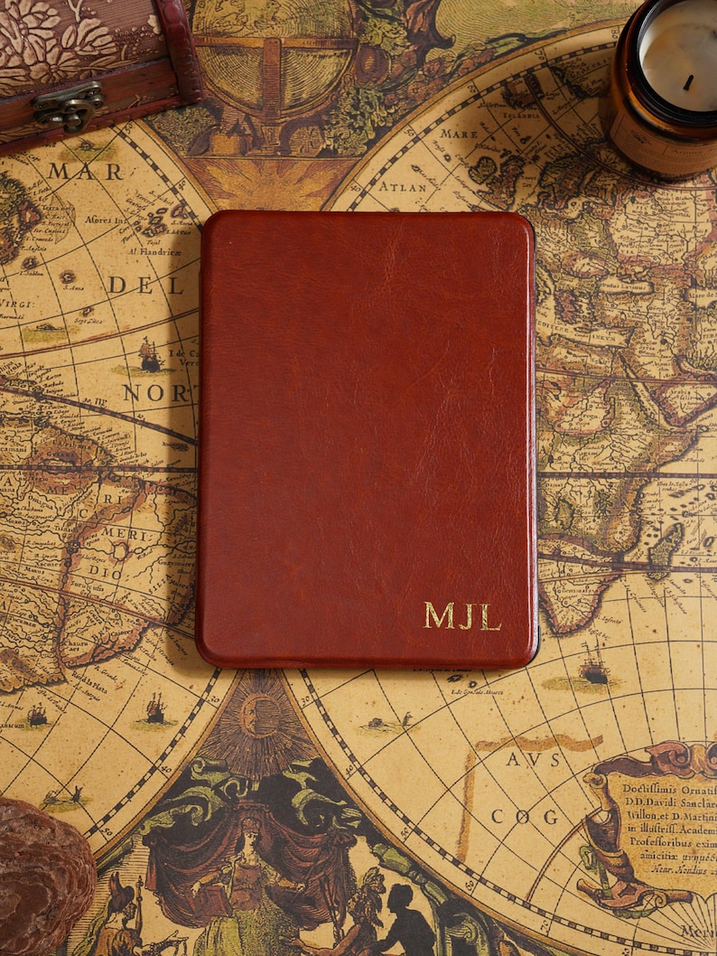 May include: A rectangular, reddish-brown leather journal with rounded corners. The initials "MJL" are embossed in gold on the lower right corner. The journal rests on a vintage map.