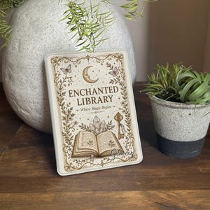 May include: A rectangular metal sign featuring a cream background with gold-toned decorative borders. The text reads ENCHANTED LIBRARY Where Magic Begins. Illustrations include a crescent moon, butterflies, crystals, an open book, and a key.