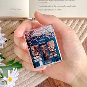 May include: A rectangular, clear acrylic book shop magnet. The magnet features a detailed illustration of a blue bookstore facade with pink cherry blossoms and the text "Book Shop". The image is held in a hand.
