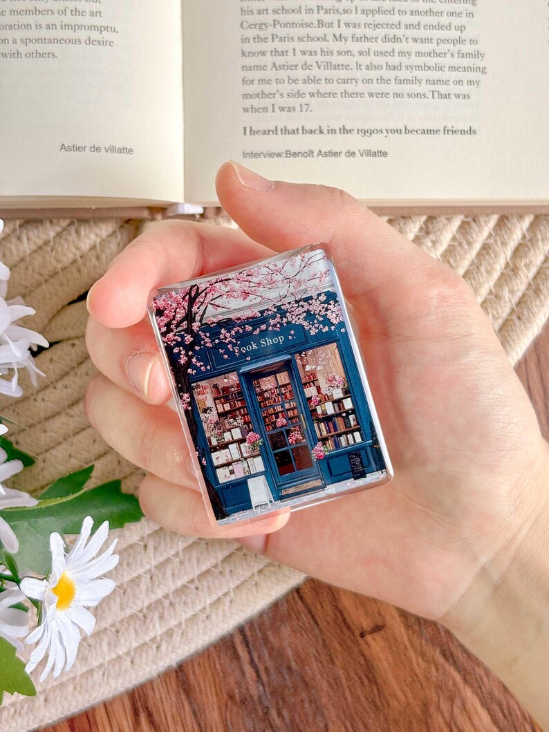 May include: A rectangular, clear acrylic book shop magnet. The magnet features a detailed illustration of a blue bookstore facade with pink cherry blossoms and the text "Book Shop". The image is held in a hand.