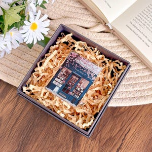 May include: A small, rectangular gift box with a gray exterior and filled with brown paper shreds. Inside, a clear acrylic block features a detailed illustration of a bookstore with the text "Book Shop" and cherry blossoms. The box rests on a woven surface.