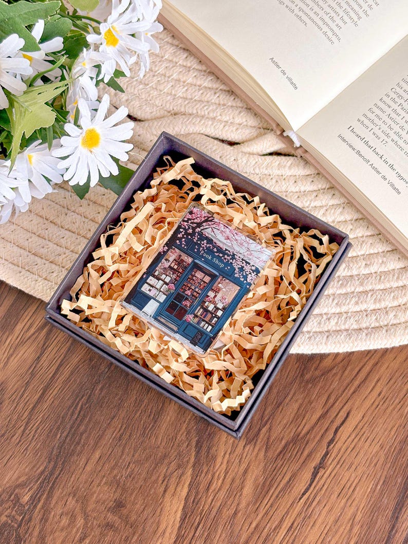 May include: A small, rectangular gift box with a gray exterior and filled with brown paper shreds. Inside, a clear acrylic block features a detailed illustration of a bookstore with the text "Book Shop" and cherry blossoms. The box rests on a woven surface.