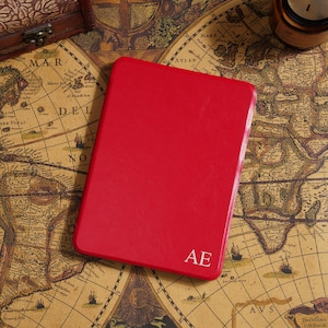 May include: A bright red leather-bound journal with the initials "AE" in white. The journal is rectangular and sits on a vintage world map. A small wooden box and a candle are also visible.