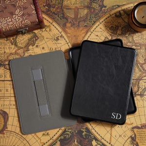 Puede incluir: Dos fundas para tabletas sobre un mapa antiguo. Una es gris con un soporte, la otra es negra con las iniciales "SD". Una pequeña caja de madera y una vela están cerca. El mapa tiene texto en latín.