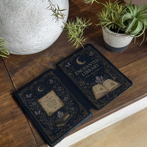 May include: A black e-reader case featuring a gold and purple celestial design with an open book, crystals, butterflies, and the text "ENCHANTED LIBRARY - Where Magic Begins" on the right side and a quote on the left side with floral accents.
