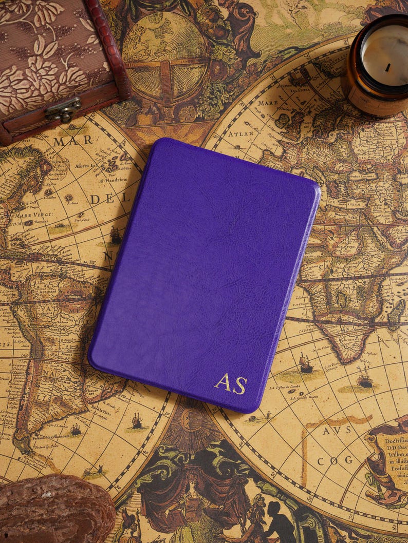 May include: A purple leather-bound journal with the gold initials "AS" on the cover. The journal is rectangular and sits on a vintage map. A small wooden box and a candle are also visible.