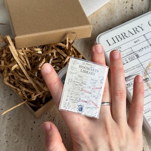 May include: A rectangular ring featuring a miniature Hogwarts Library checkout card design with black text on a white background, including the words Hogwarts Library and Overdue, set in a silver-toned metal band.