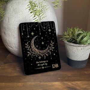 May include: A black rectangular item with a celestial design featuring a crescent moon, stars, and gold decorative elements. The text "She belongs to the night sky" is visible, along with the initials "DB". It is displayed on a wooden surface.