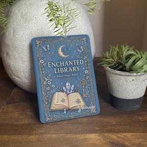May include: A rectangular blue sign featuring the text "ENCHANTED LIBRARY Where Magic Begins" in gold lettering. The design includes illustrations of a crescent moon, butterflies, a golden key, crystals, and an open book with handwritten text.