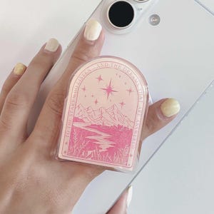 以下が含まれることがあります： 山、星、そして「TO THE STARS WHO LISTEN AND THE DREAMS THAT ARE ANSWERED」というテキストが特徴のピンク色の携帯電話グリップ。 グリップは透明な携帯電話ケースに取り付けられています。