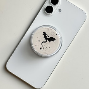 May include: A white smartphone with a round, clear phone grip attached. The grip features a black dragon silhouette with stars and clouds on a beige background. The phone has two black camera lenses.