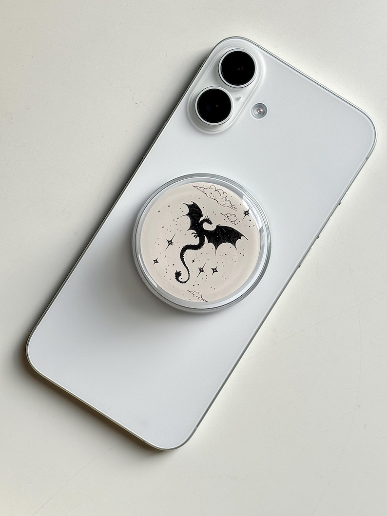 May include: A white smartphone with a round, clear phone grip attached. The grip features a black dragon silhouette with stars and clouds on a beige background. The phone has two black camera lenses.