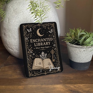 May include: A rectangular black sign featuring gold-toned illustrations of a crescent moon, butterflies, a key, and an open book with crystals. The text reads "ENCHANTED LIBRARY" and "Where Magic Begins" with the name "Sherry" in white script.