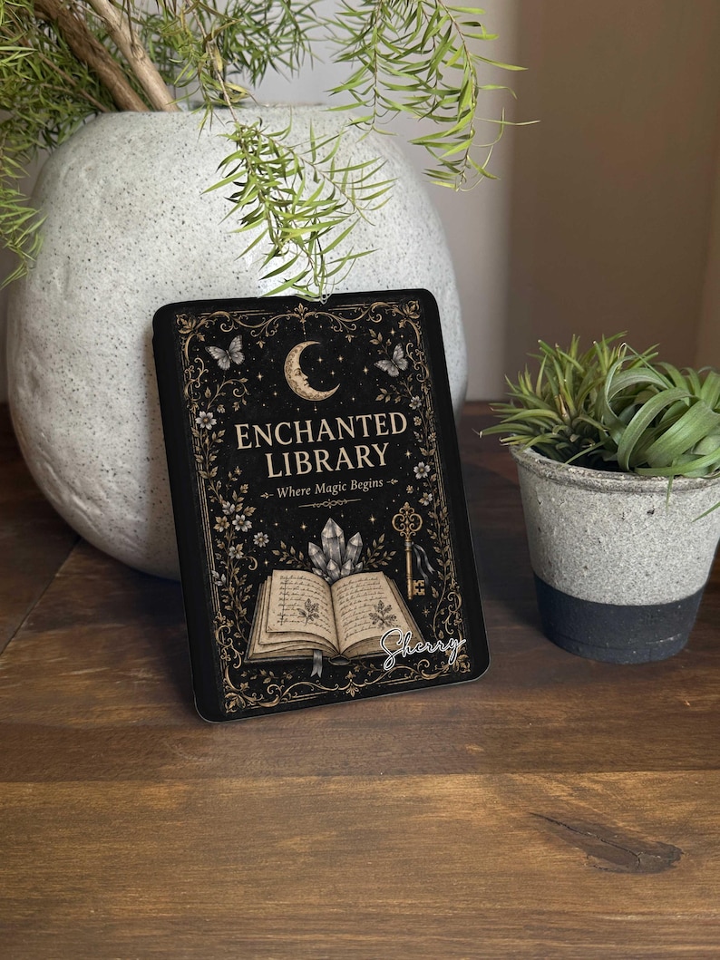 May include: A rectangular black sign featuring gold-toned illustrations of a crescent moon, butterflies, a key, and an open book with crystals. The text reads "ENCHANTED LIBRARY" and "Where Magic Begins" with the name "Sherry" in white script.