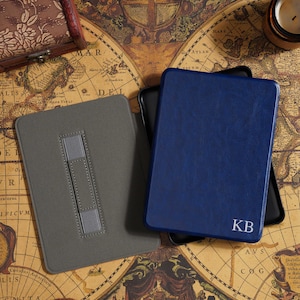 May include: A gray and a navy blue leather e-reader case with a personalized monogram. The gray case has a stand. The navy blue case has the initials "KB" in white. The cases are on a vintage map.