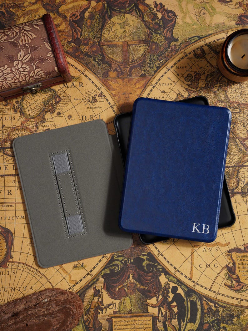 May include: A gray and a navy blue leather e-reader case with a personalized monogram. The gray case has a stand. The navy blue case has the initials "KB" in white. The cases are on a vintage map.