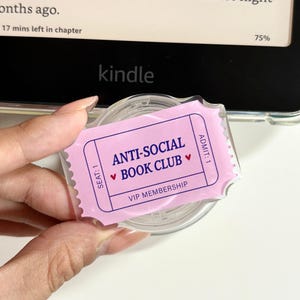 以下が含まれることがあります： 透明な円形のプラスチック製アイテムで、薄紫色のチケット型のステッカーが貼られています。ステッカーには「ANTI-SOCIAL BOOK CLUB」という文字と小さな赤いハート、さらに「SEAT: 1」「ADMIT: 1」「VIP MEMBERSHIP」という文字が濃い青色で印刷されています。