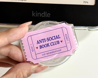 May include: A clear, circular plastic item featuring a light purple ticket-shaped sticker. The sticker displays the text ANTI-SOCIAL BOOK CLUB with small red hearts, along with the words SEAT: 1, ADMIT: 1, and VIP MEMBERSHIP in dark blue ink.