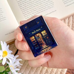 May include: A rectangular, dark blue pin featuring a detailed illustration of a bookstore at night. The pin has the words "Book Store" in gold lettering above the store's entrance, with illuminated windows and a starry sky.