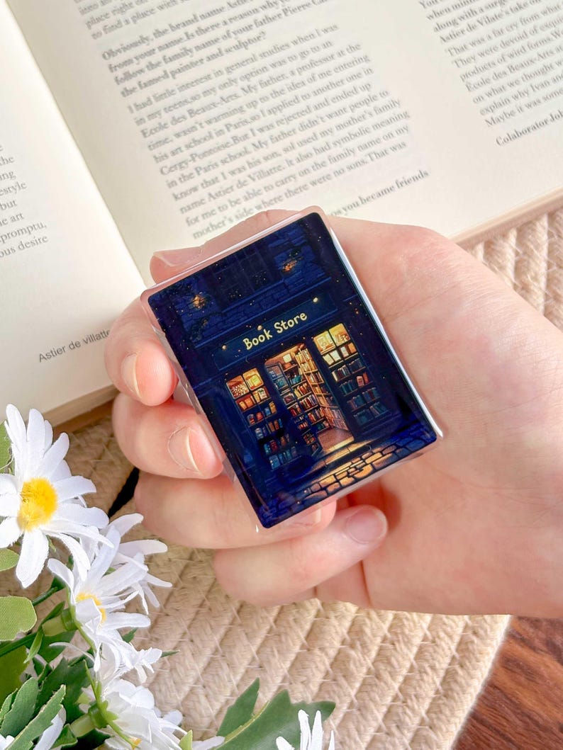 May include: A rectangular, dark blue pin featuring a detailed illustration of a bookstore at night. The pin has the words "Book Store" in gold lettering above the store's entrance, with illuminated windows and a starry sky.