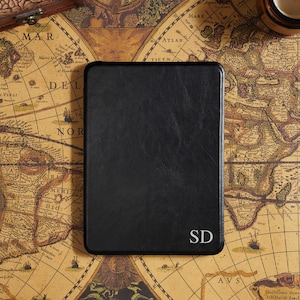 May include: A black leather-bound journal with rounded corners, featuring the initials "SD" in the lower right corner. The journal rests on a vintage map, with a small candle and a decorative box in the background.