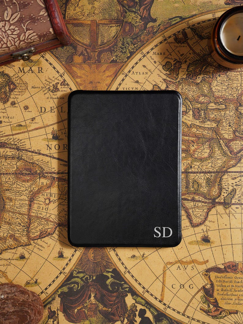 May include: A black leather-bound journal with rounded corners, featuring the initials "SD" in the lower right corner. The journal rests on a vintage map, with a small candle and a decorative box in the background.