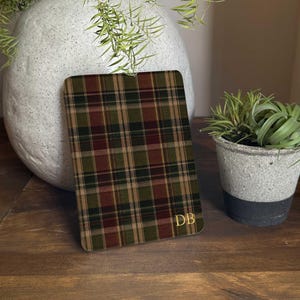 Puede incluir: Un objeto plano rectangular con un patrón de cuadros en tonos verde oscuro, marrón y beige. La superficie tiene esquinas redondeadas y muestra las letras mayúsculas doradas DB en la esquina inferior derecha, en fuente serif.