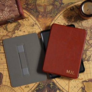 May include: A collection of leather-bound journals and cases in shades of gray, black, and red-brown, with gold-colored initials on one. The items are arranged on a vintage world map, suggesting a travel or writing theme.