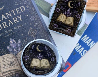 May include: A rectangular and a circular phone grip featuring a dark illustration of an open book, purple crystals, butterflies, and a crescent moon against a starry background. The design includes text on the book pages and a gold-toned border.