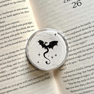 May include: A round, clear acrylic pop socket featuring a black dragon design with stars and clouds on a white background. The dragon is centered, with its wings spread. The pop socket is set against an open book.