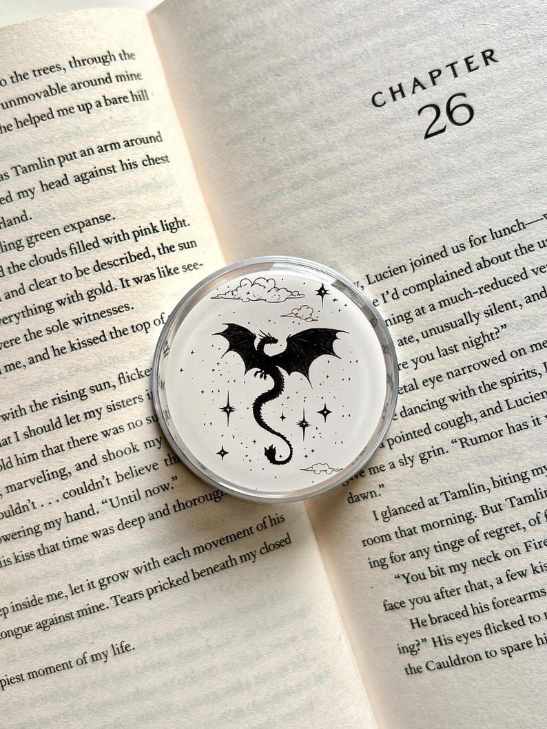 May include: A round, clear acrylic pop socket featuring a black dragon design with stars and clouds on a white background. The dragon is centered, with its wings spread. The pop socket is set against an open book.