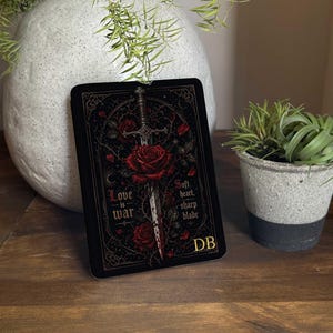 Puede incluir: Una tarjeta rectangular negra con un diseño gótico que presenta una espada que atraviesa una rosa roja, rodeada de rosas y el texto "Love is war" y "Soft heart, sharp blade". Las iniciales "DB" están en la esquina inferior derecha.
