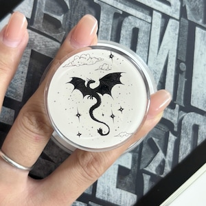 May include: A round, clear container with a black dragon illustration. The dragon has outstretched wings and a long tail, surrounded by stars and clouds. The container is held in a hand with a silver ring.