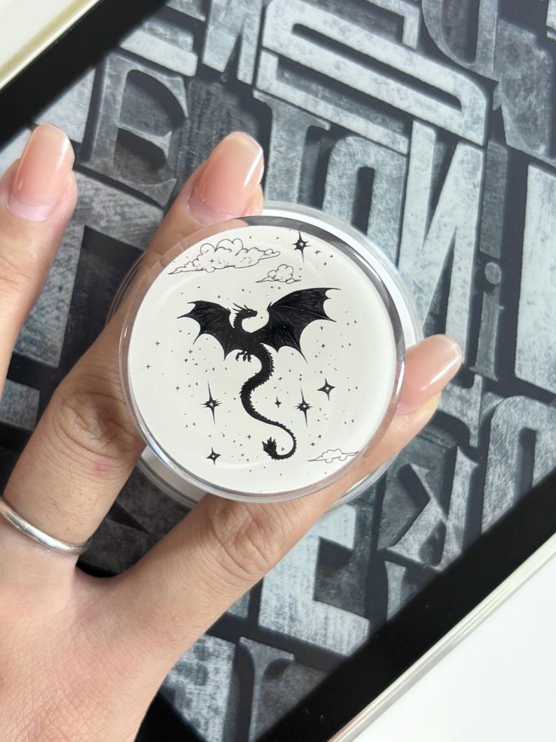May include: A round, clear container with a black dragon illustration. The dragon has outstretched wings and a long tail, surrounded by stars and clouds. The container is held in a hand with a silver ring.