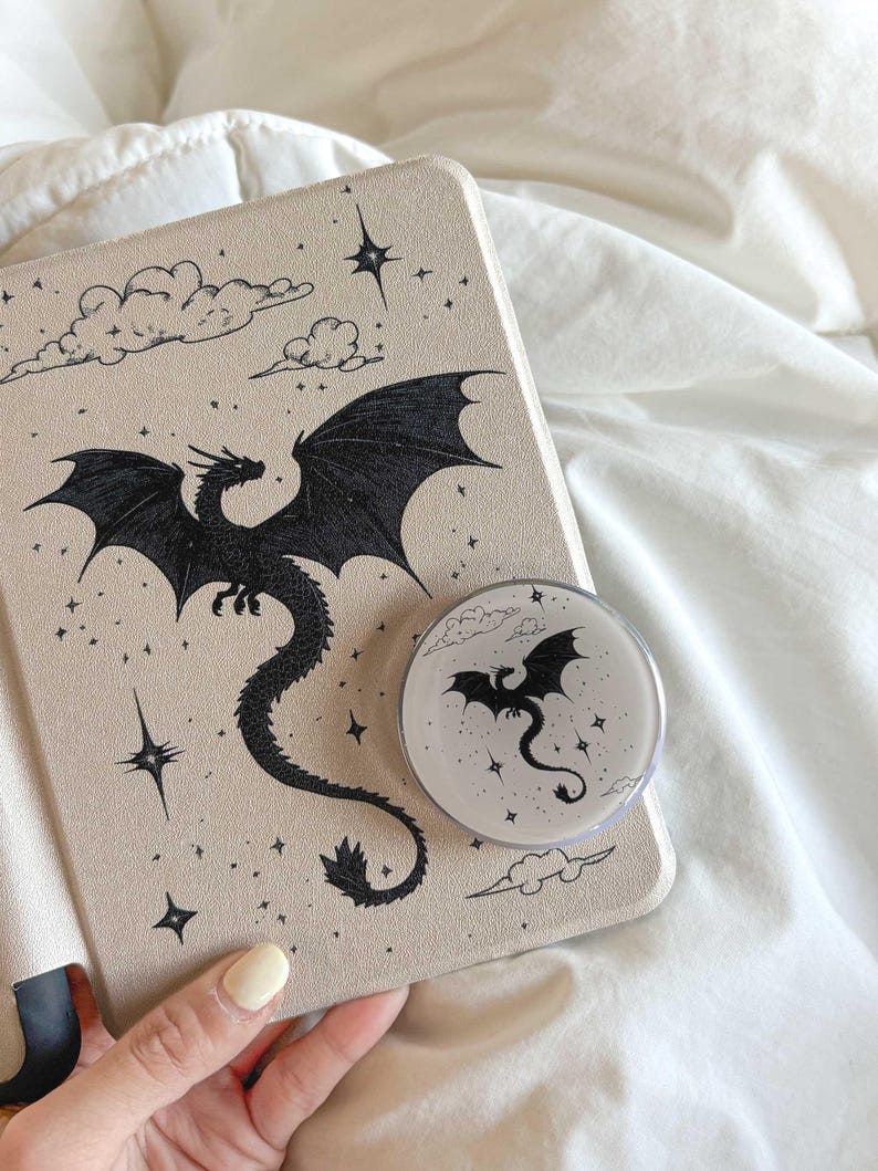 May include: A beige tablet case and a round phone grip featuring a black dragon design. The dragon is depicted with outstretched wings, surrounded by stars and clouds. The phone grip mirrors the dragon design on the case.