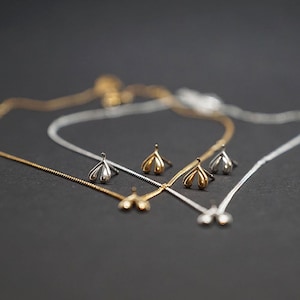 Op de afbeelding: Close-up van twee delicate kettingen en bijpassende oorknopjes. De ene ketting- en oorbellen set is goudkleurig, de andere zilverkleurig. De sieraden hebben een bloemmotief tegen een donkere achtergrond.