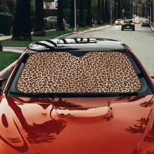 Può includere: Parasole per auto con motivo leopardato, visibile attraverso il parabrezza di un'auto rossa. Il parasole è beige con macchie marroni. L'auto è parcheggiata in una strada con palme.