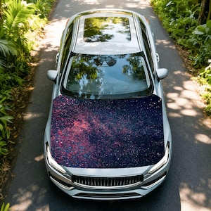 May include: A silver car with a panoramic sunroof and a hood featuring a galaxy design in shades of purple, red, and blue. The car is driving on a paved road surrounded by lush green foliage.